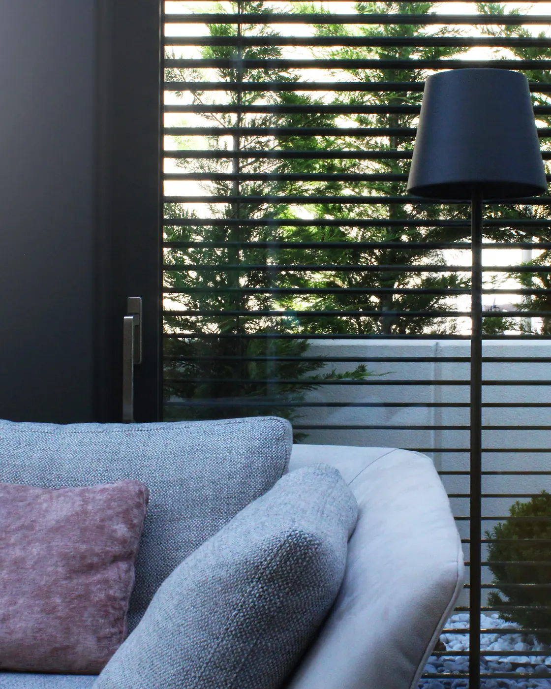 Interno di un giardino d'inverno: divano chiaro e lampada da terra davanti a una parete vetrata con frangisole a lame orizzontali, verde oltre il vetro.