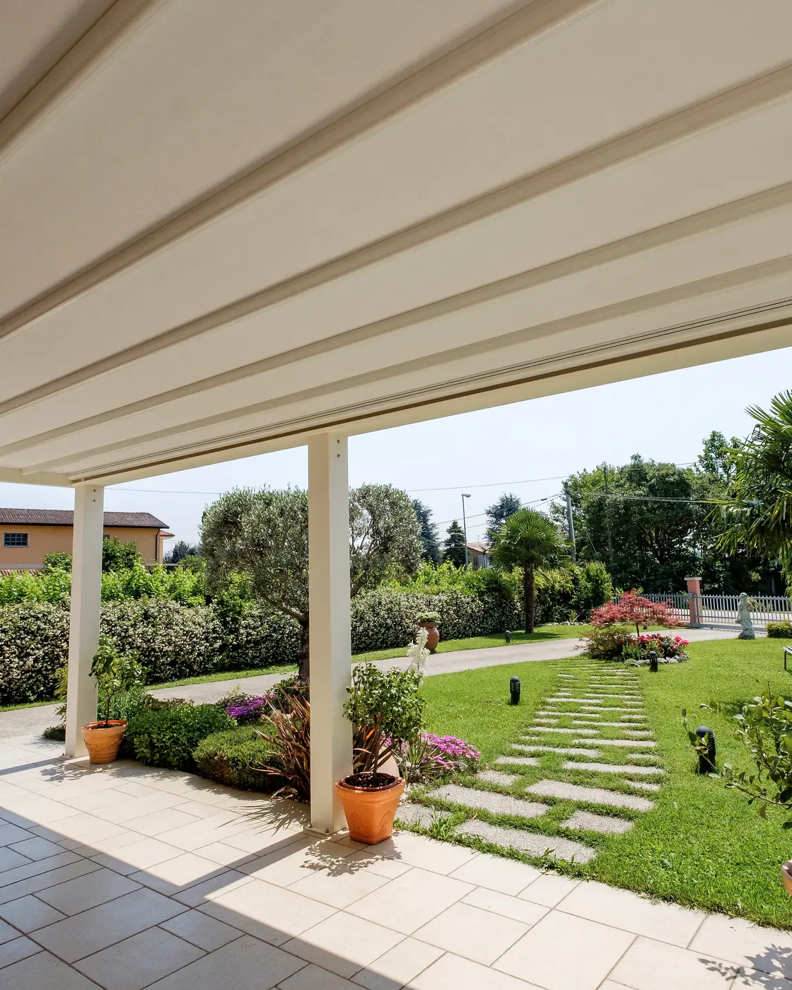 Pergola addossata a una casa vista da sotto: copertura a lame chiare, montanti allineati, giardino e camminamento in pietra oltre il bordo.