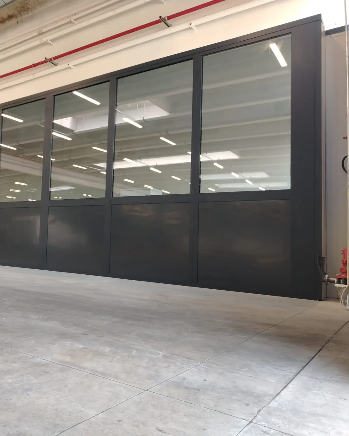 Box ufficio dentro un capannone: parete vetrata continua con profili scuri lunga tutta la campata, pavimento industriale e travi a vista sopra.