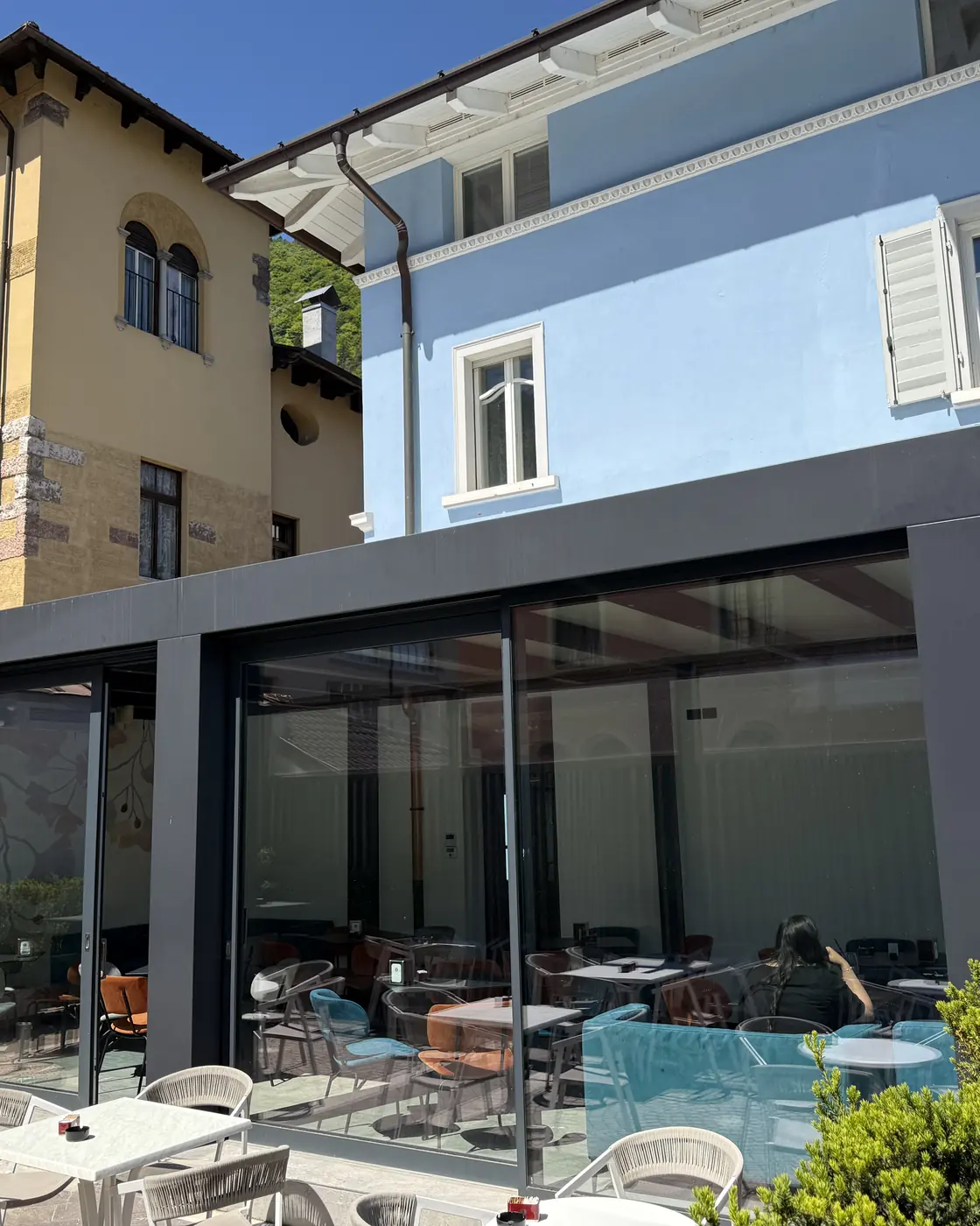Veranda vetrata addossata a un edificio azzurro: profili scuri a filo, vetri a tutta altezza, tavolini apparecchiati davanti e dentro.