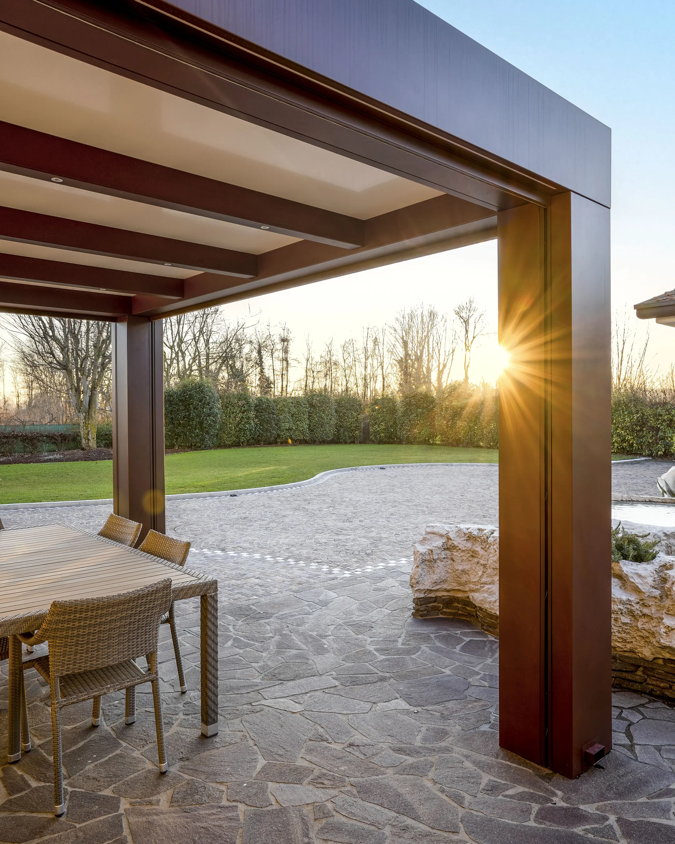 Vista da sotto una pergola bioclimatica: struttura in alluminio brunito, lame del tetto, il sole basso che filtra fra i montanti su un giardino.